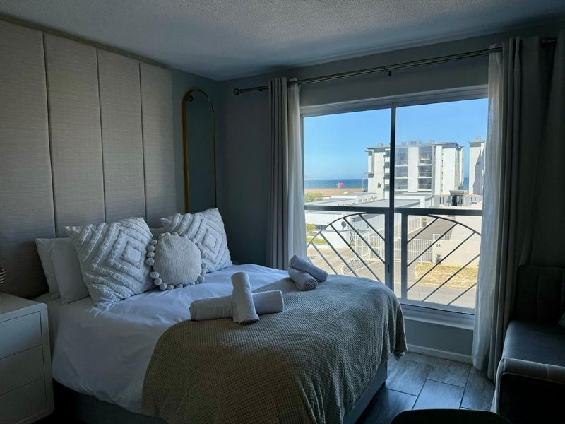 A well-maintained bedroom featuring floor-to-ceiling upholstered wall paneling, tiled flooring, and a sliding glass door opening to a balcony with coastal views.
