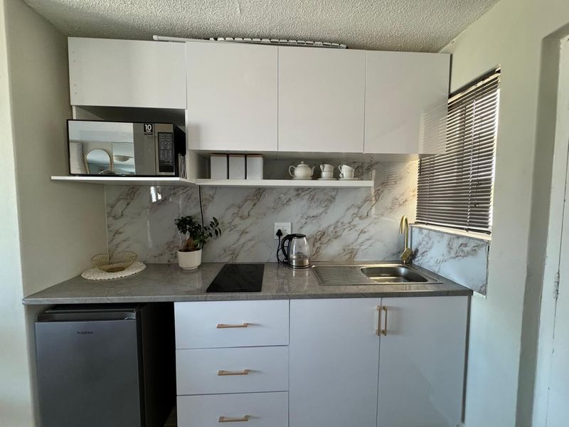 Compact modern kitchen featuring white cabinetry, marble-look splashback, stone-look countertop, integrated electric hob, and a brass faucet with a window view.