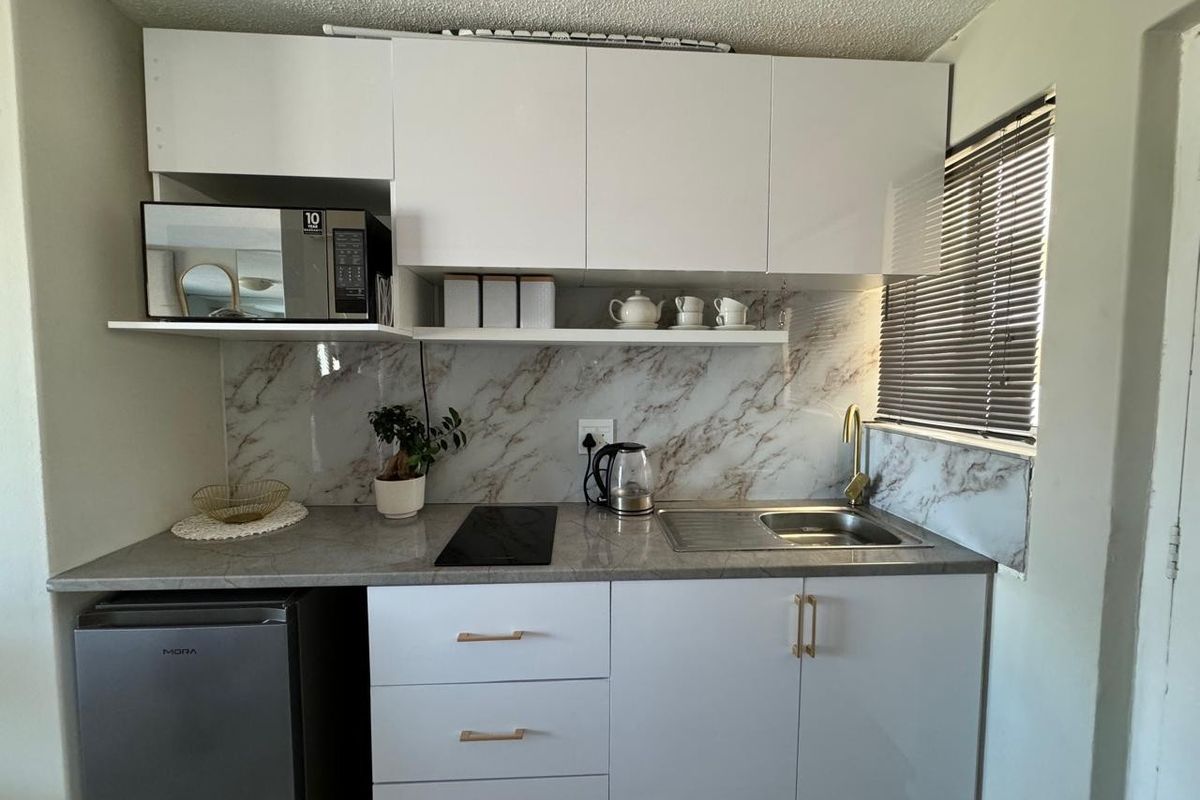 Compact modern kitchen featuring white cabinetry, marble-look splashback, stone-look countertop, integrated electric hob, and a brass faucet with a window view.