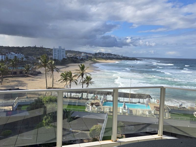 Stunning elevated coastal view overlooking the beach, ocean, and communal pool area from a private balcony with a modern glass balustrade.