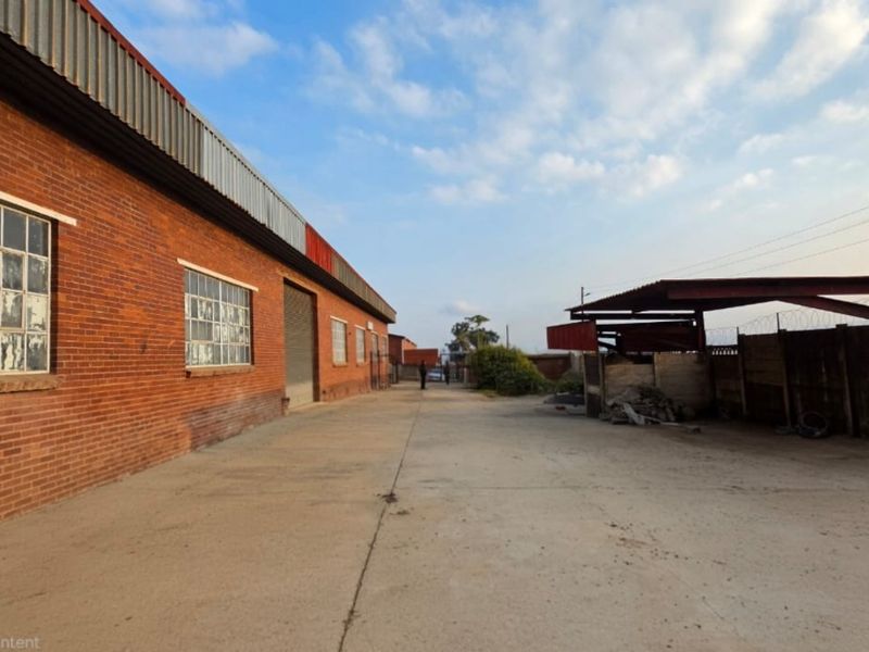 Rear industrial yard featuring a brick warehouse building with metal roofing, steel-framed windows, a roller door, and a covered outdoor storage or loading area.