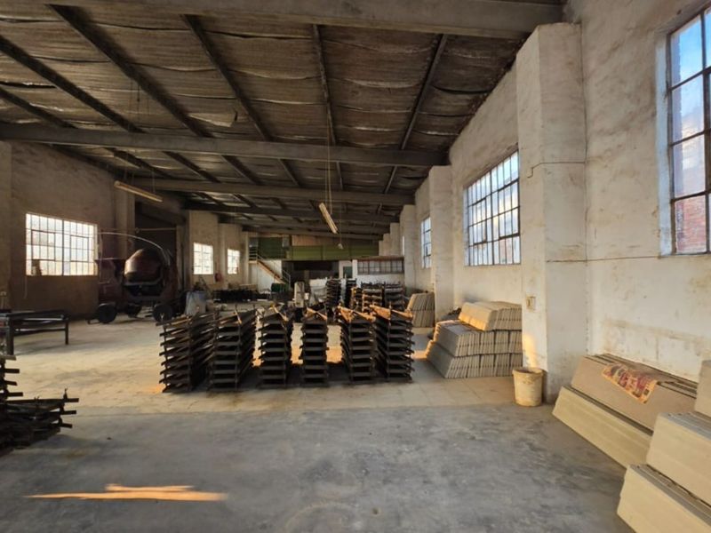 Spacious industrial warehouse area featuring high ceilings with exposed steel trusses, concrete flooring, and large steel-framed windows providing natural light.