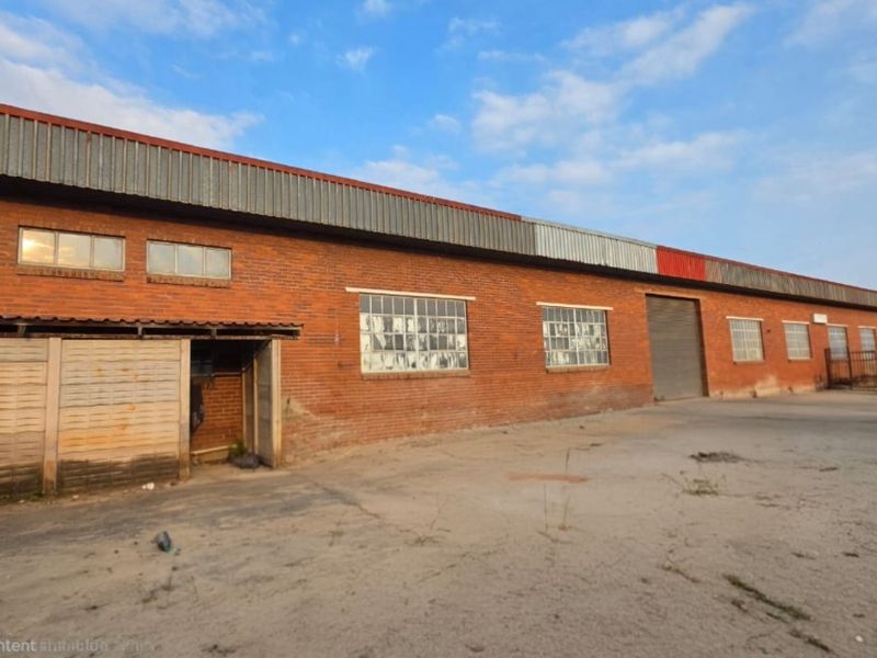Industrial warehouse exterior featuring a red brick facade, metal roofing, roller shutter access, and a large concrete yard area.