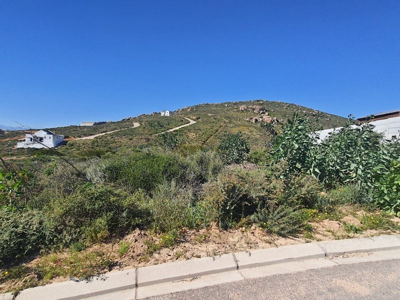A vacant residential plot featuring natural vegetation, a concrete curb, and a paved road frontage with surrounding hillside views.