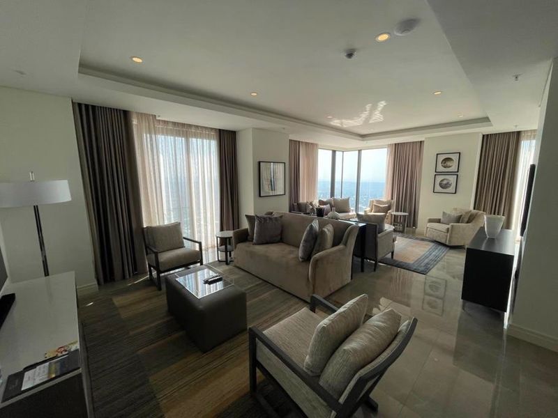 A spacious, modern living room featuring high ceilings with recessed lighting, polished stone floors, and expansive floor-to-ceiling windows offering panoramic views.