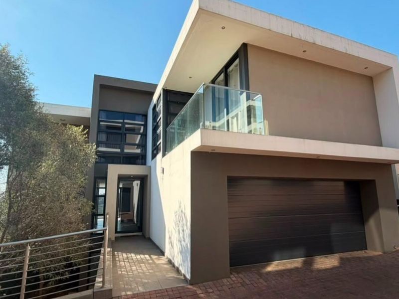 Modern double-story residential facade featuring a glass balcony, sectional garage door, and paved driveway entrance.
