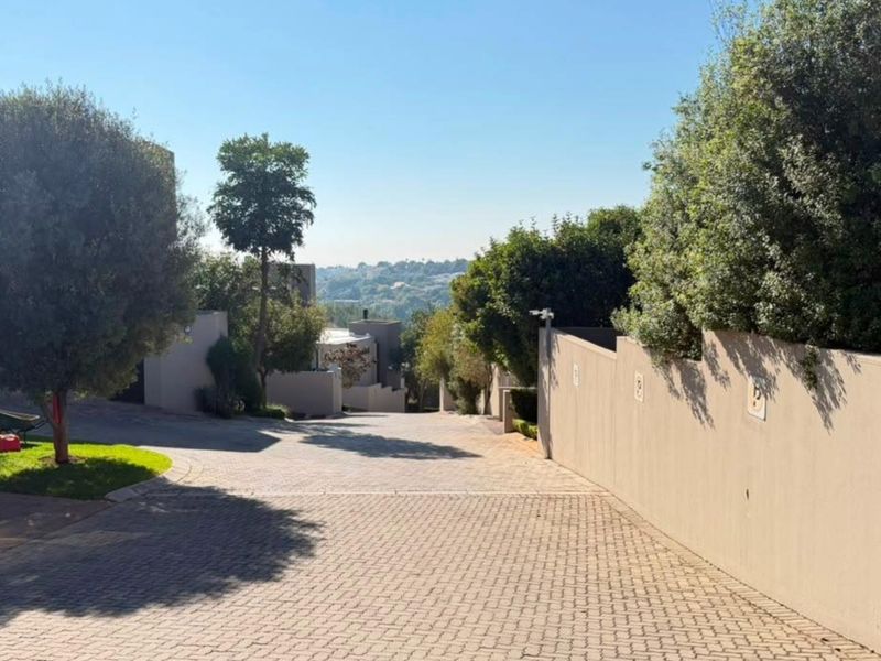 A view looking down a paved residential driveway towards the property entrance, featuring boundary walls and established trees in a suburban setting.