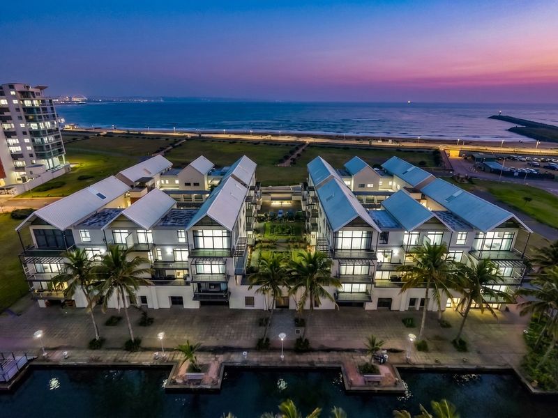 Aerial view of a modern apartment complex featuring pitched roofs, private balconies, and a central landscaped courtyard, situated near the coastline.