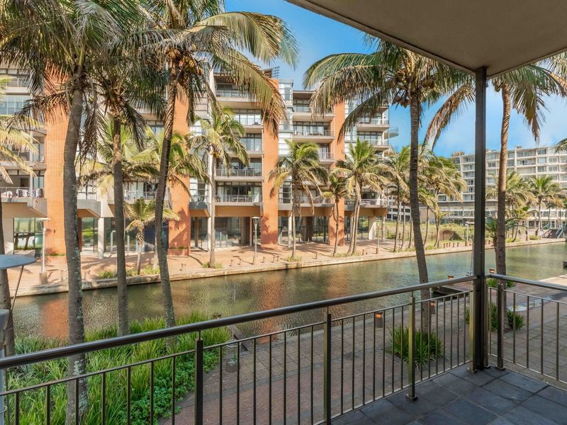 A spacious covered balcony featuring a metal balustrade, offering scenic views of the canal and surrounding apartment complex.