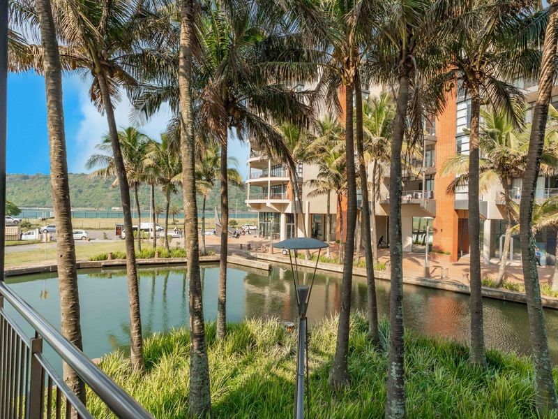 A scenic view from a balcony overlooking a tranquil water feature, landscaped gardens with palm trees, and the surrounding apartment complex architecture.