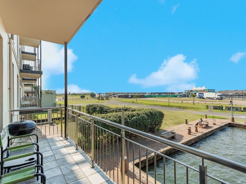 A covered balcony featuring tiled flooring and a stainless steel balustrade, offering an open view of the waterfront and surrounding landscape.