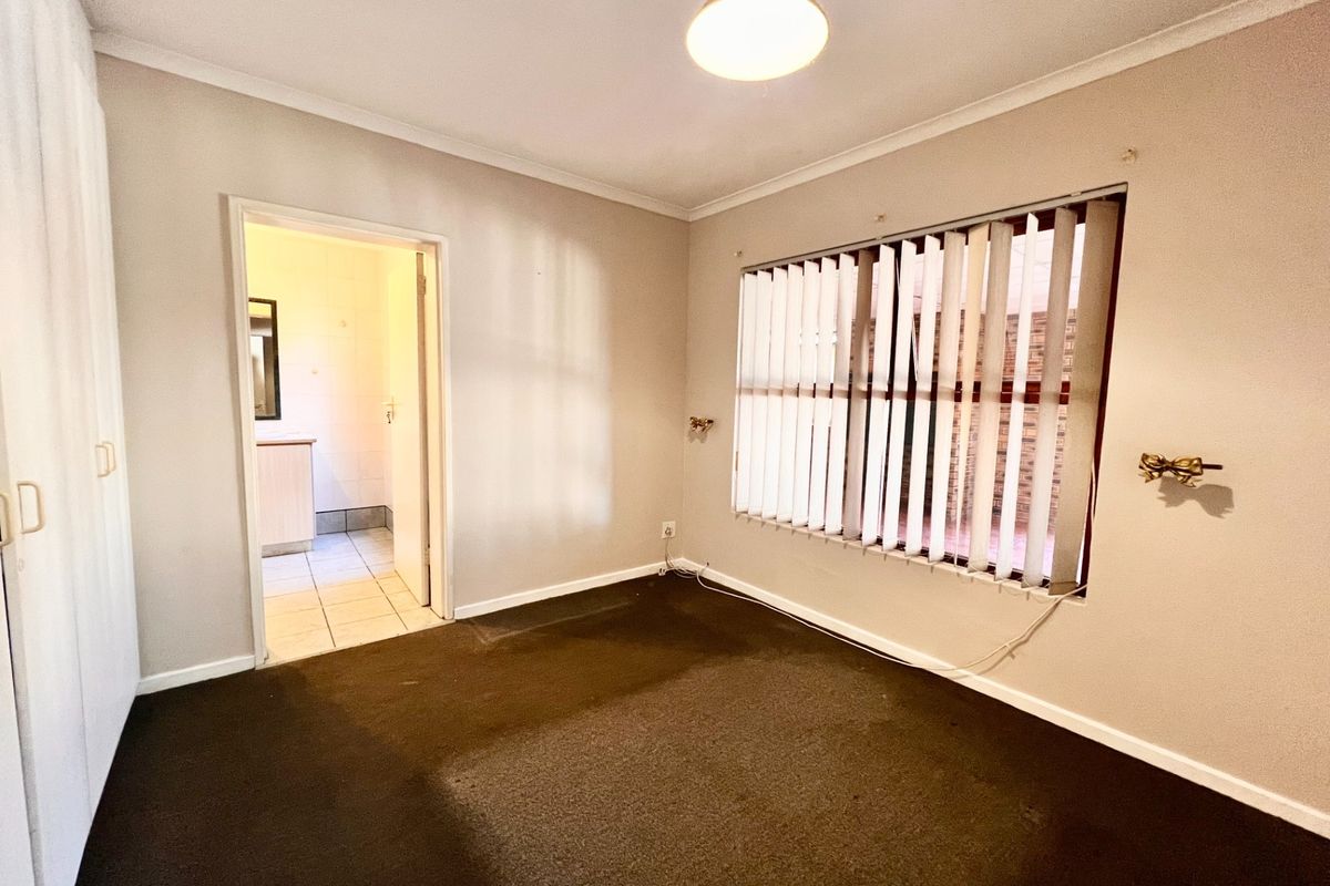 An empty bedroom featuring built-in cupboards, carpet flooring, and vertical blinds, with an adjoining en-suite bathroom visible through the doorway.