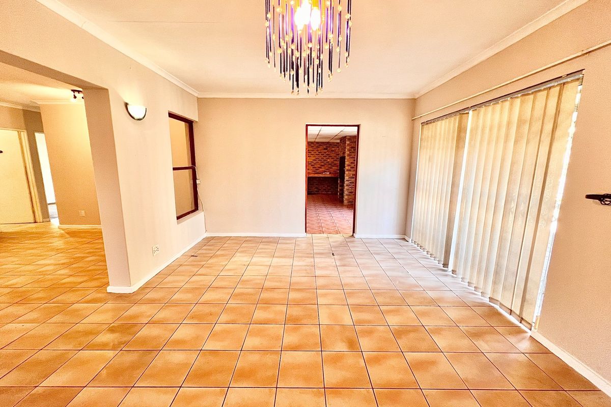 An empty, open-plan living area featuring tiled flooring, a decorative chandelier, and large windows with vertical blinds, leading through to an adjacent room.