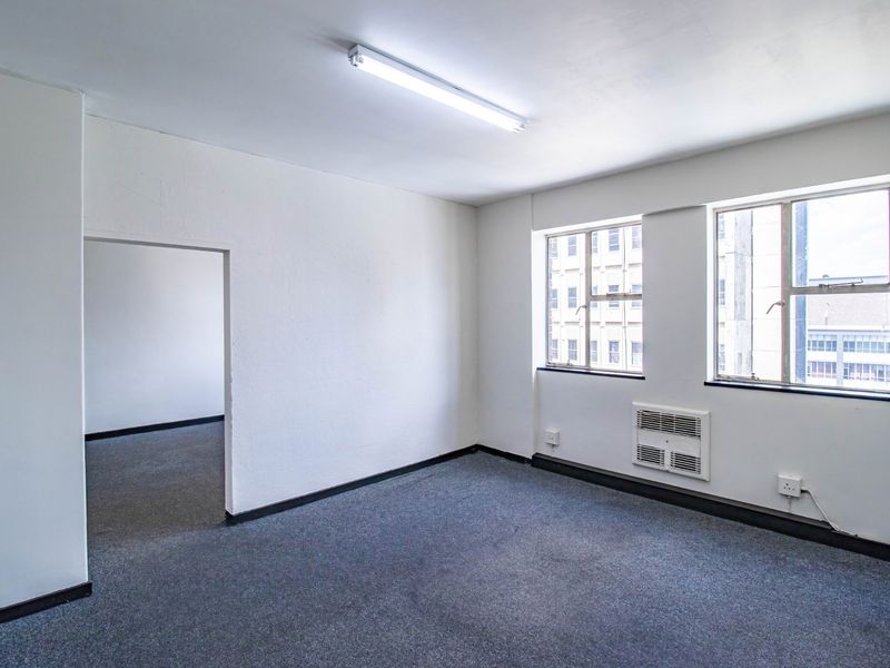 A vacant, open-plan office space featuring grey carpeting, built-in air conditioning, and natural light from large windows.
