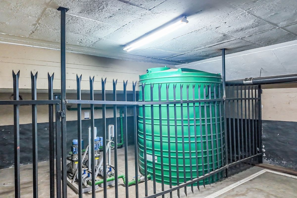 Utility area featuring a large green water storage tank and associated pump system, secured within a fenced enclosure with exposed concrete ceiling.