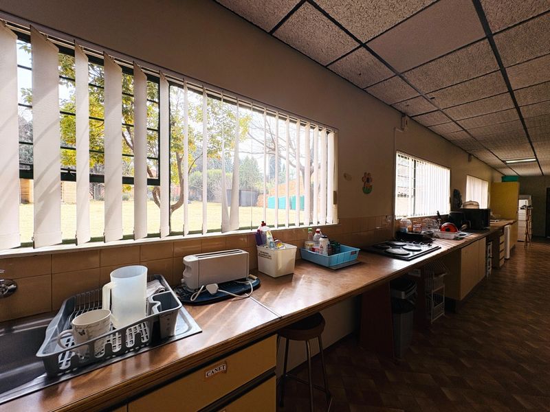 A long, functional kitchen featuring extensive laminate counter space, a built-in hob, and large windows with vertical blinds overlooking the garden.