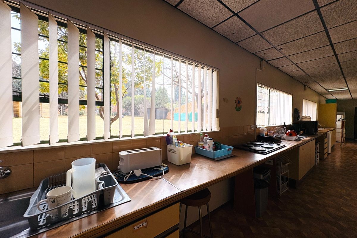 A long, functional kitchen featuring extensive laminate counter space, a built-in hob, and large windows with vertical blinds overlooking the garden.