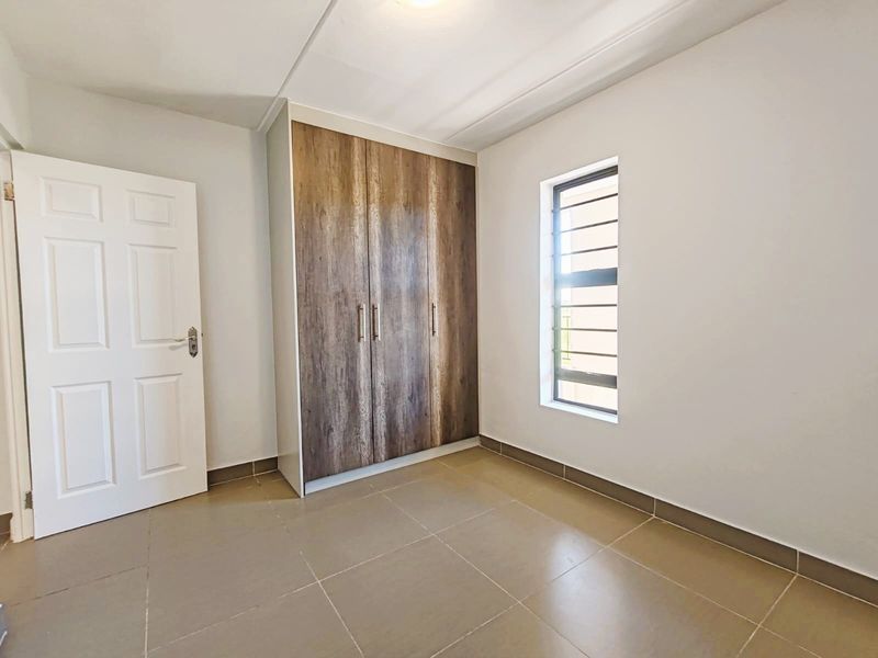 A clean, empty bedroom featuring modern wood-finish built-in cupboards, neutral tiled flooring, and a louvered window providing natural light.