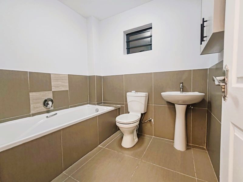 A clean, modern bathroom featuring a built-in bathtub, pedestal basin, toilet, and neutral-toned wall and floor tiling.