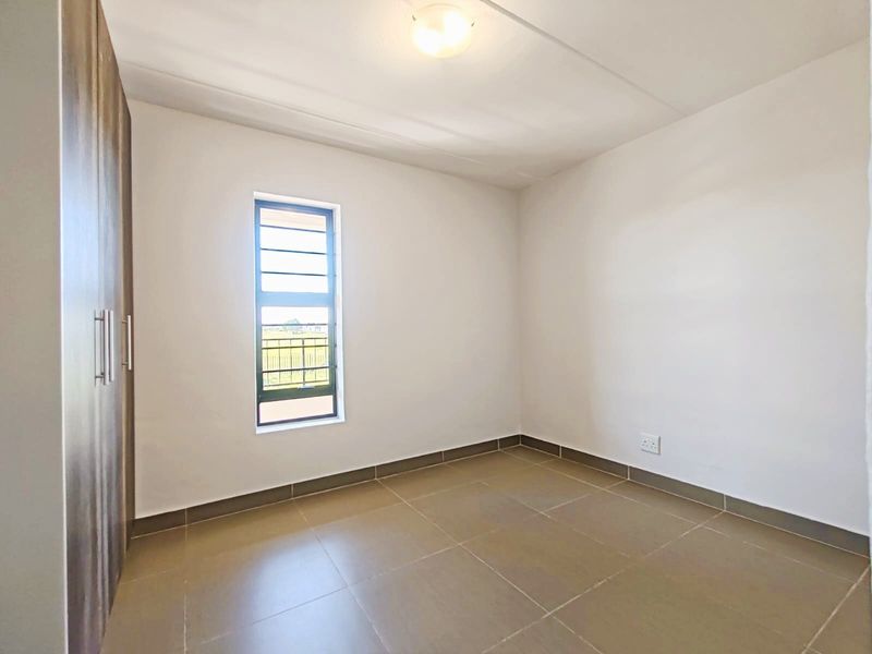 A clean, empty bedroom featuring neutral tiled flooring, built-in cupboards, and a vertical window providing natural light.