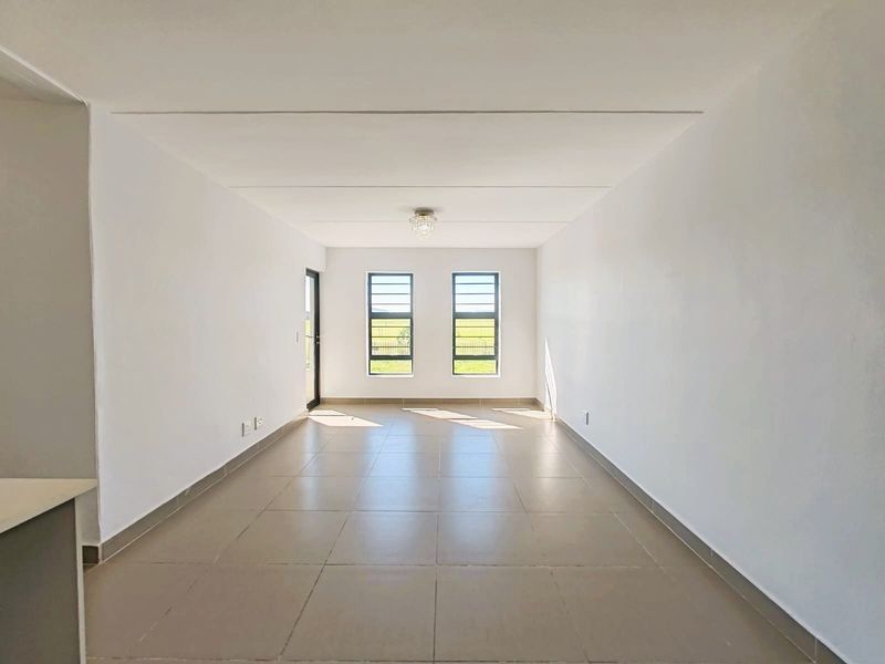 A bright, empty open-plan living area featuring neutral tiled flooring, clean white walls, and large aluminium-framed windows allowing for ample natural light.