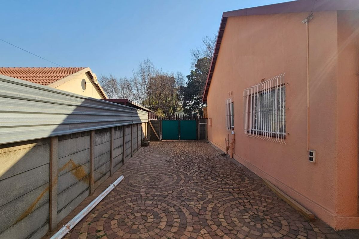 A side exterior view of the property featuring a paved driveway, secure boundary walling, and security gates.