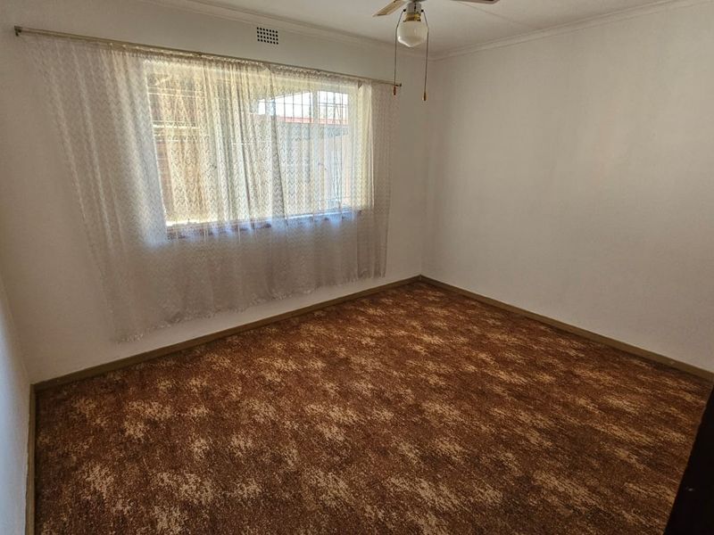 An empty bedroom featuring brown patterned carpet flooring, a ceiling fan, and a window fitted with security burglar bars.
