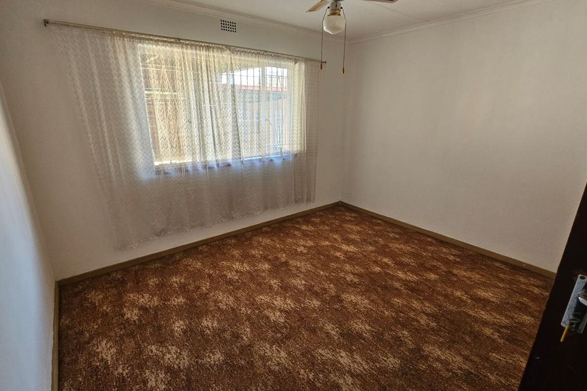 An empty bedroom featuring brown patterned carpet flooring, a ceiling fan, and a window fitted with security burglar bars.
