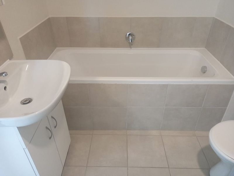 A clean, neutral-toned bathroom featuring a built-in bathtub, vanity cabinet with basin, and tiled flooring and walls.