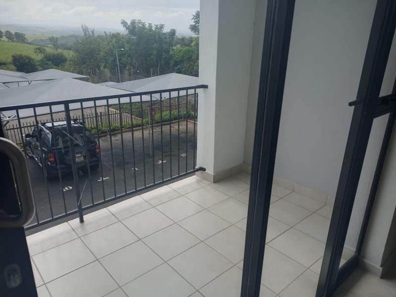 A clean, tiled balcony featuring a metal balustrade and sliding glass door access, overlooking the parking area and surrounding landscape.