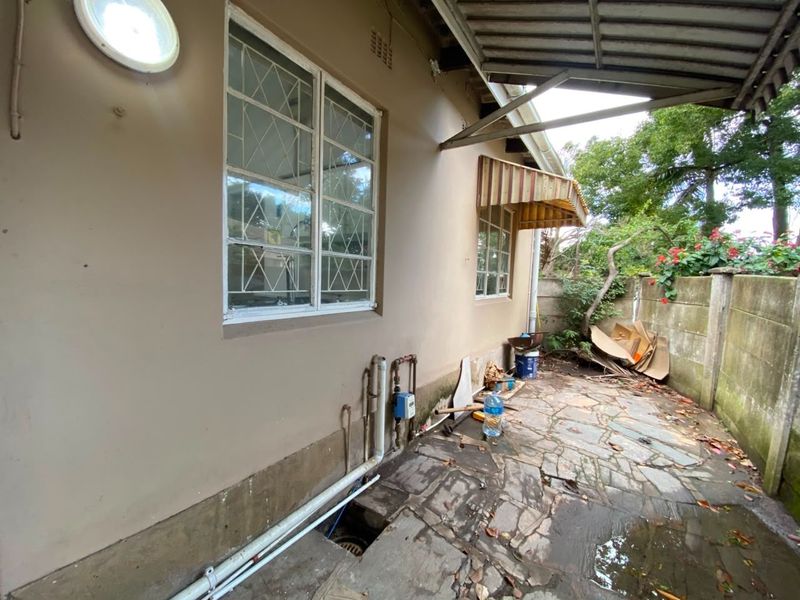 Rear exterior area of the cottage featuring stone paving, security-barred windows, and a small awning, requiring maintenance and clearing of debris.