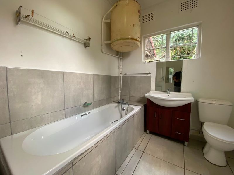 A functional bathroom featuring a bathtub, vanity unit, and toilet, with tiled finishes and an exposed electric water heater.