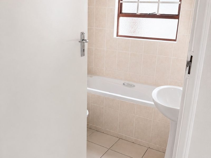 A functional bathroom featuring a bathtub, pedestal basin, and neutral wall and floor tiling, viewed through an open doorway.