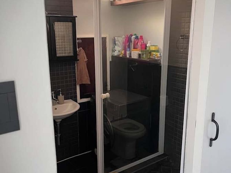 A compact bathroom featuring a glass-enclosed shower, wall-mounted basin, and dark mosaic tiling, providing a functional space with built-in storage.
