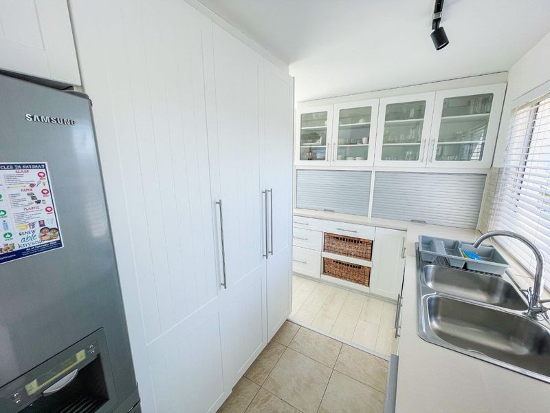 A well-maintained scullery featuring white built-in cabinetry, glass-fronted wall units, double stainless steel sink, and tiled flooring.
