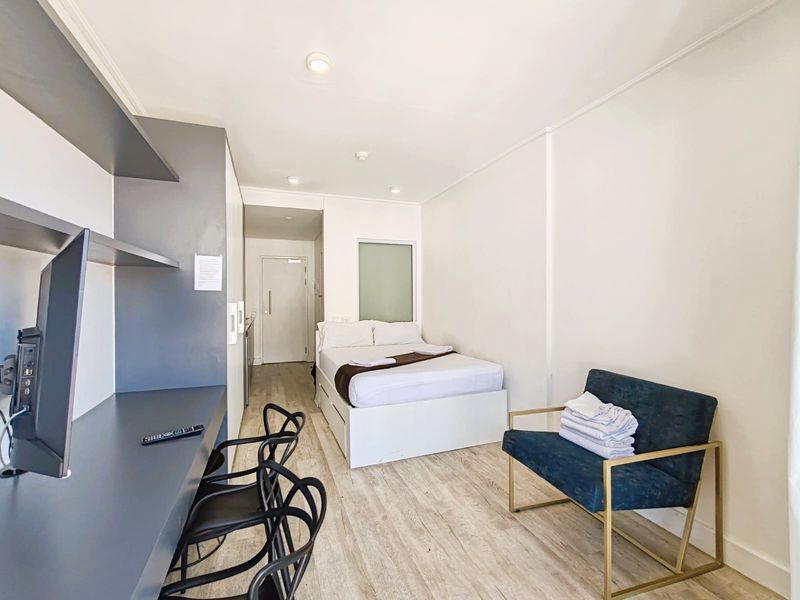 A compact bedroom featuring light laminate flooring, built-in desk and shelving unit, recessed ceiling lights, and a built-in wardrobe.