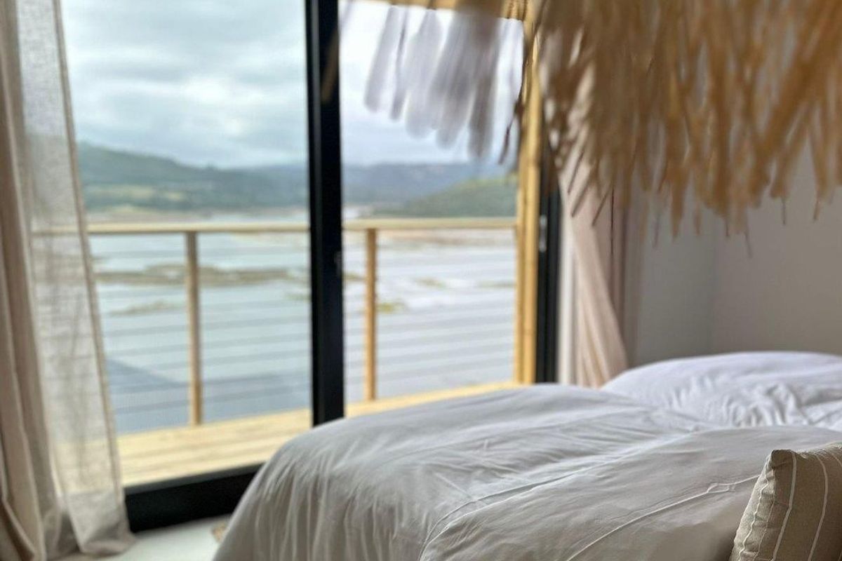 A bright bedroom featuring large sliding glass doors that open onto a wooden deck with scenic water views.