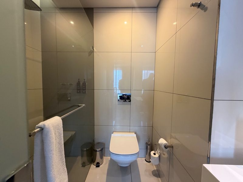 Modern bathroom featuring a wall-mounted toilet, glass shower screen with rain shower head, and neutral floor-to-ceiling tiling.