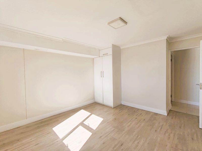 A bright, empty bedroom featuring light-toned laminate flooring, neutral walls, and a functional built-in cupboard with overhead storage.