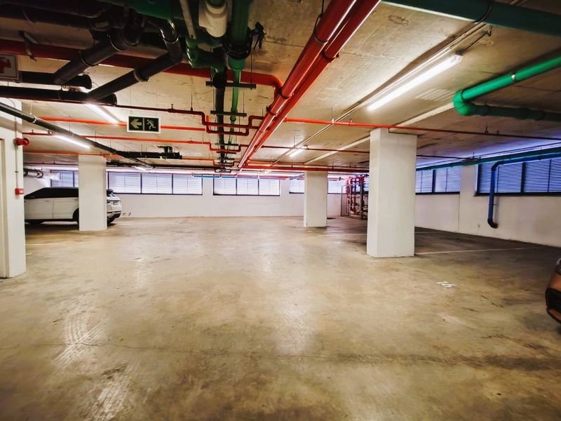 A spacious, well-lit underground parking garage featuring exposed industrial piping, concrete columns, and marked parking bays.