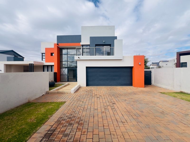 Modern double-story residence featuring a bold orange and grey facade, double garage, paved driveway, and a prominent glass-fronted entrance with an upper-level balcony.