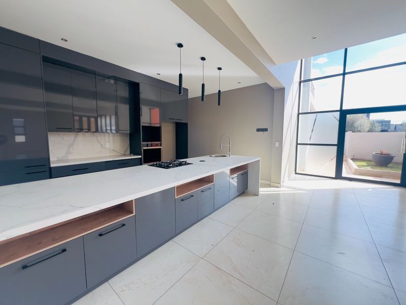 Modern kitchen featuring sleek grey cabinetry, quartz countertops, integrated appliances, and expansive floor-to-ceiling windows providing natural light and garden access.