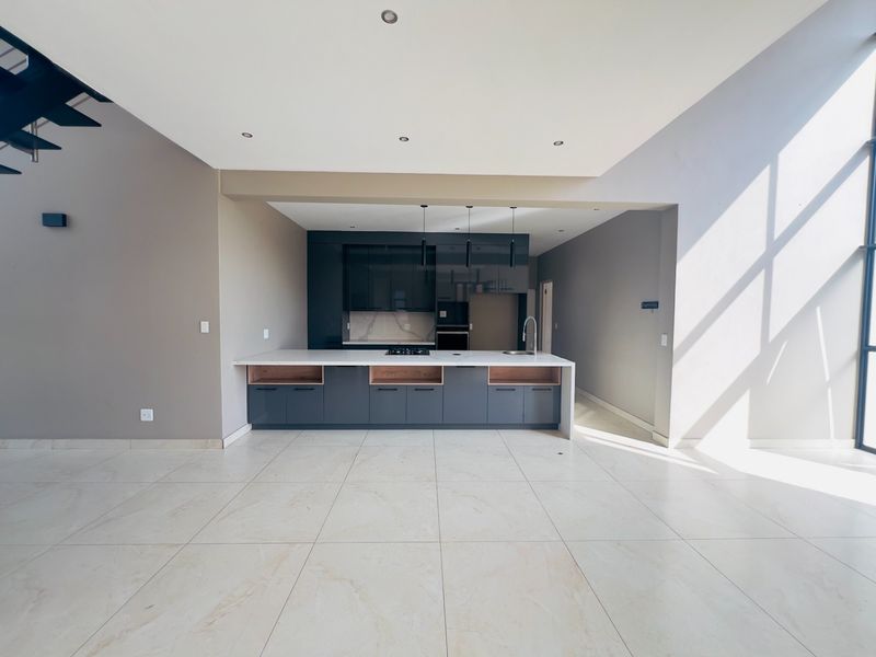 Modern open-plan kitchen featuring sleek dark cabinetry, a central island with a gas hob, and light-toned tiled flooring in a spacious, sunlit living area.