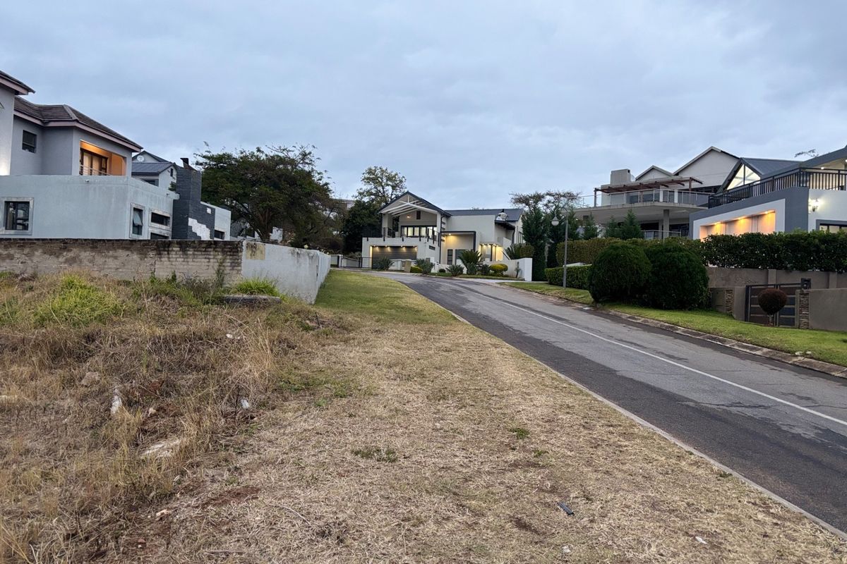 A street-level view of a residential neighborhood featuring an asphalt road, paved driveway, and surrounding established homes with boundary walls.