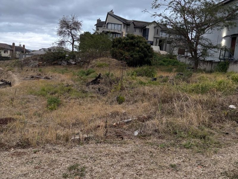 A vacant residential plot featuring sloped terrain and natural vegetation, situated in an established neighborhood with surrounding residential properties.