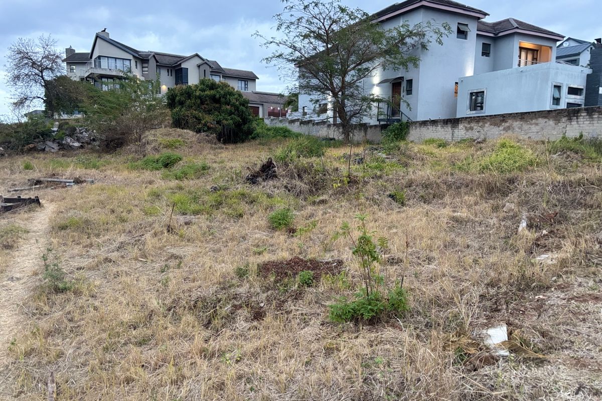 A vacant residential plot featuring uneven, overgrown terrain with an existing boundary wall, situated in an established neighborhood.