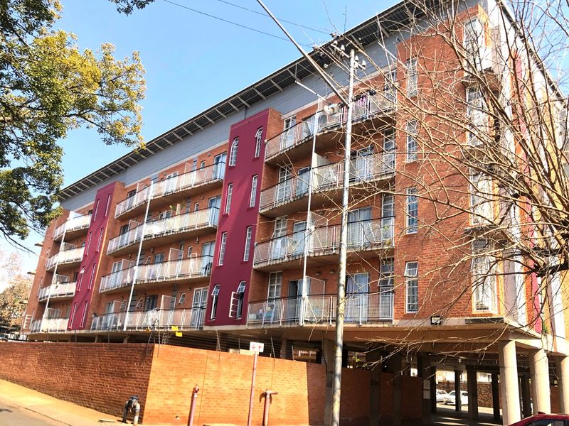 A multi-story apartment building featuring a red brick facade, private balconies with metal railings, and integrated covered parking facilities.