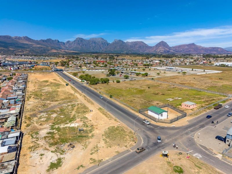 An aerial view of a commercial development site featuring road access, perimeter fencing, and surrounding infrastructure with mountain views in the background.