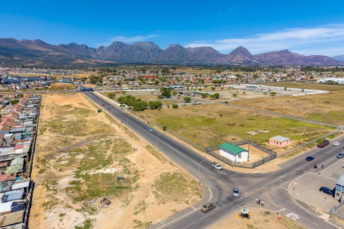 An aerial view of a commercial development site featuring road access, perimeter fencing, and surrounding infrastructure with mountain views in the background.