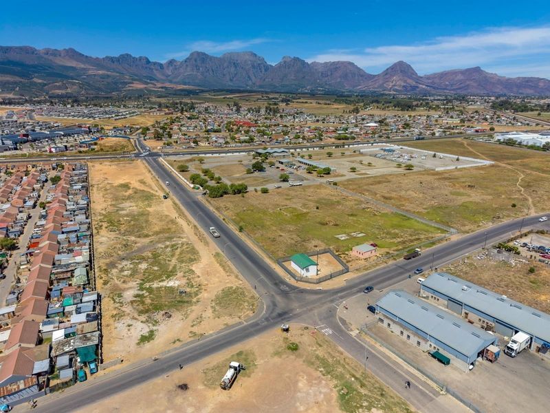 Aerial view of a commercial land plot situated at a road intersection, featuring nearby industrial warehouses and surrounding residential development.